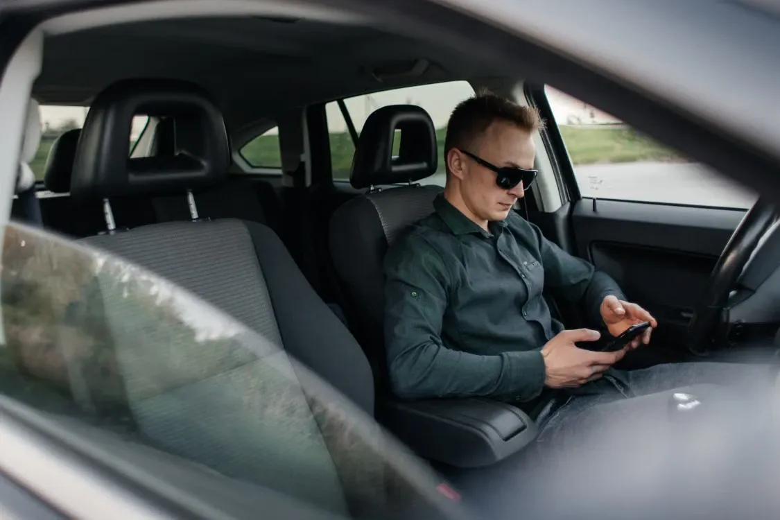 A driver in sunglasses checking his phone in the front seat of his car
