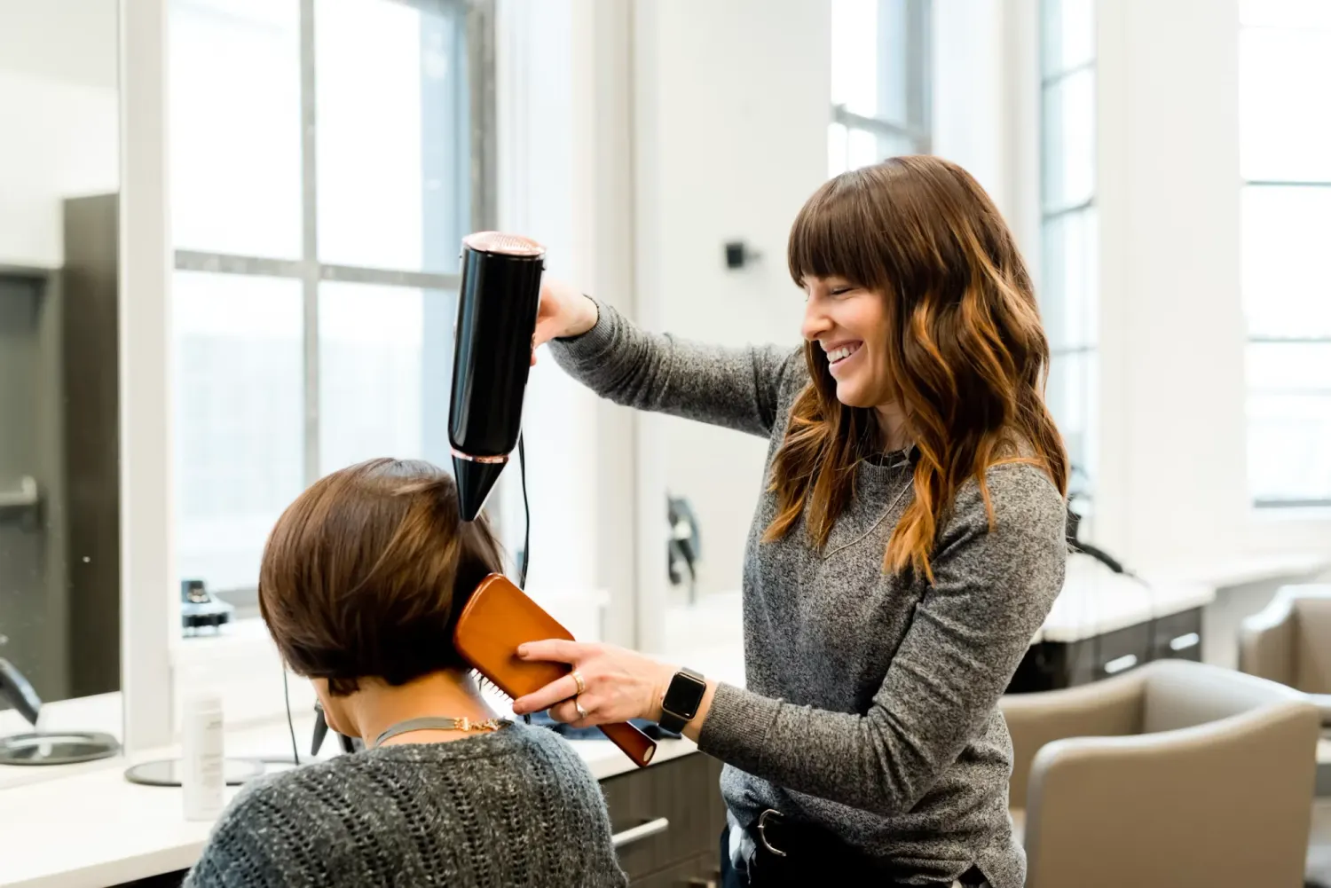 A smiling hairdresser blow-drying a client's hair in her salon
